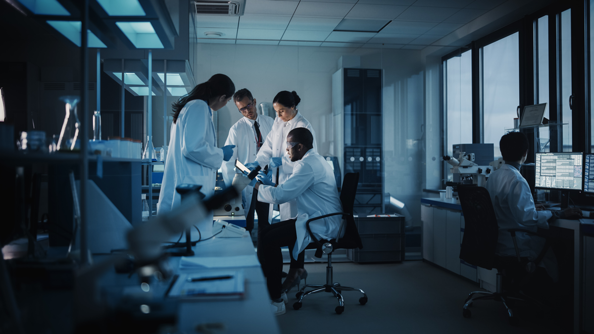 Researchers in lab coats gather around a work station.